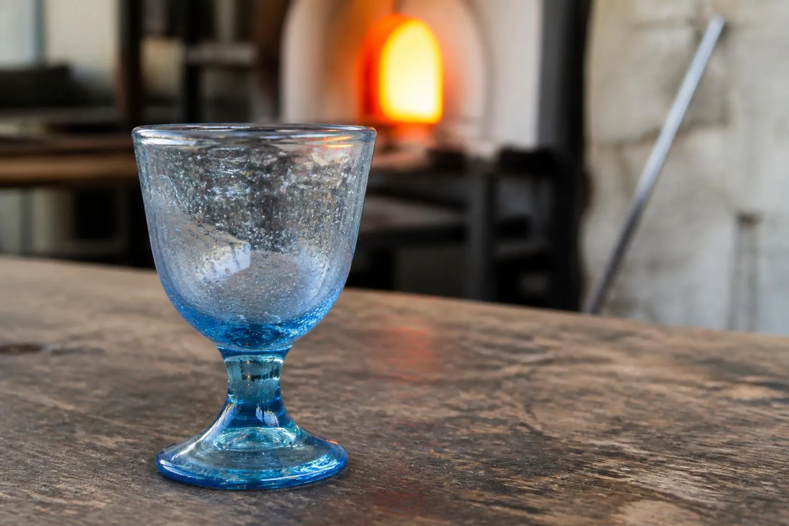 Verre à pied bullé soufflé à la bouche, teinte bleu pâle, posé sur un établi de verrier devant la lueur d'un four