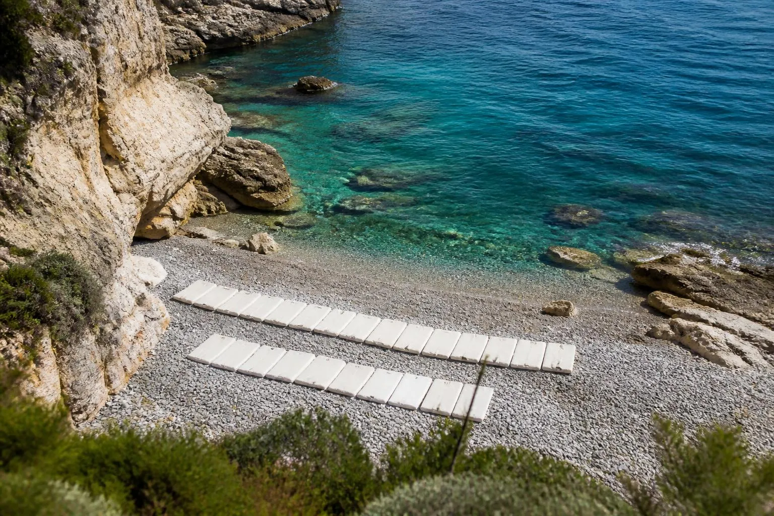 La crique de galets de la Mala vue du haut de l'escalier, la falaise, l'eau turquoise et les rangées de matelas de la plage privée